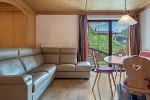 A cozy room with a leather sofa, wooden walls, a dining table with chairs, and a view of greenery and mountains through the window.