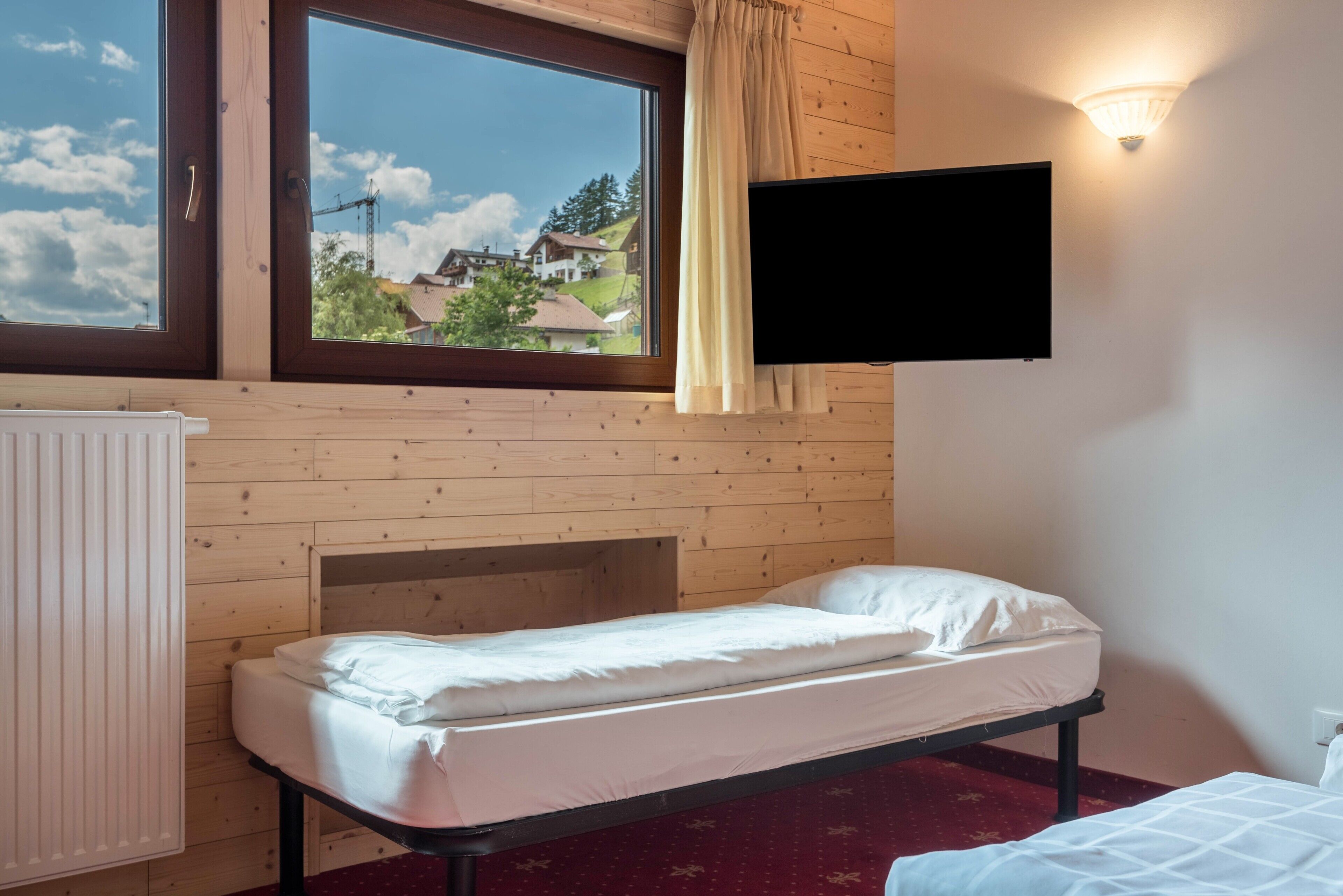 A single bed with a headboard, a television mounted on the wall, a window with curtains, and a radiator.