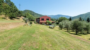 A house with a red roof, surrounded by a grassy lawn and trees, with mountains in the background. - Villa 'Rifugio Arcobaleno' with Mountain View, Heated Pool & Wi-Fi (Popiglio)
