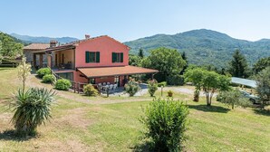 A two-story house with a red facade, a balcony, and a view of green hills. - Villa 'Rifugio Arcobaleno' with Mountain View, Heated Pool & Wi-Fi (Popiglio)