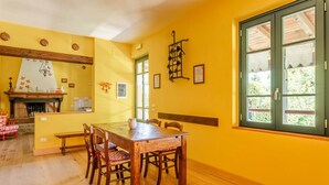 A dining area with a wooden table and chairs, a fireplace, and a window with a view of greenery. - Villa 'Rifugio Arcobaleno' with Mountain View, Heated Pool & Wi-Fi (Popiglio)