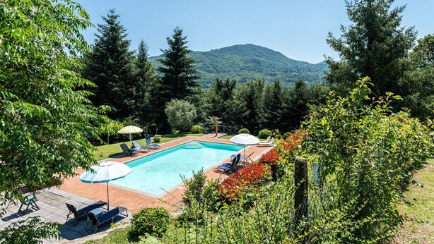A swimming pool with lounge chairs and umbrellas, surrounded by trees and a mountain in the background. - Apartment 'Alba al Rifugio Arcobaleno' with Mountain View, Shared Pool & Wi-Fi (Popiglio)