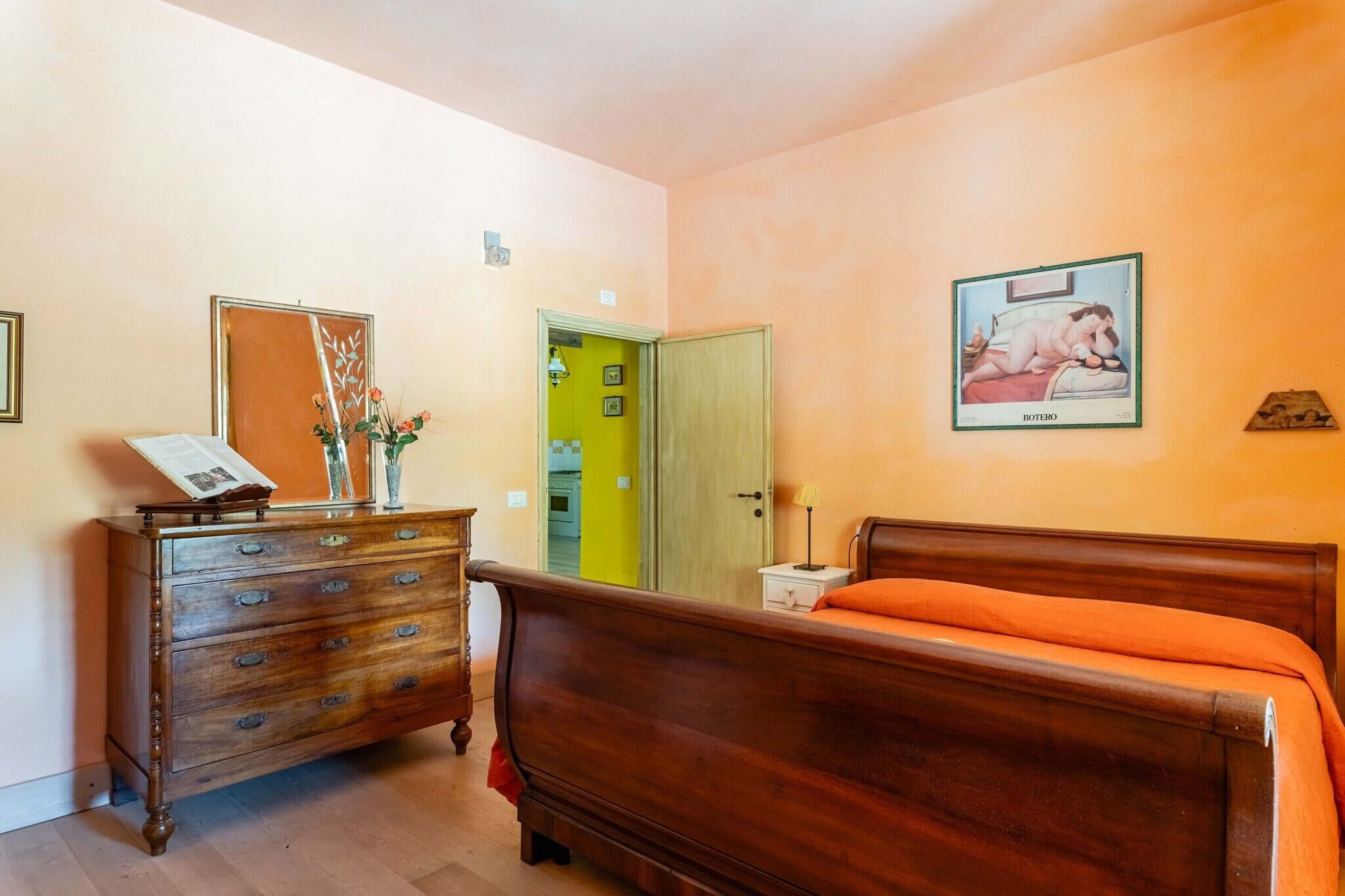 A bedroom with a wooden dresser, a bed with an orange bedspread, a green door, and a framed picture on the wall.