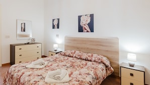 Ein Schlafzimmer mit einem hölzernen Kopfteil, einer blumigen Bettdecke, einem Nachttischchen mit einer Lampe und einer Kommode mit Schubladen.