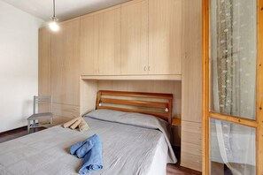 A bedroom with a bed, a chair, wooden wardrobe, and a window with lace curtains.