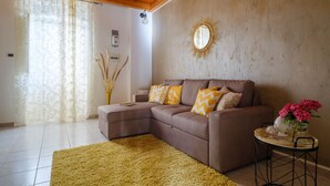 A living room with a sectional sofa, a round mirror, a small table with flowers, and a large window with sheer curtains.