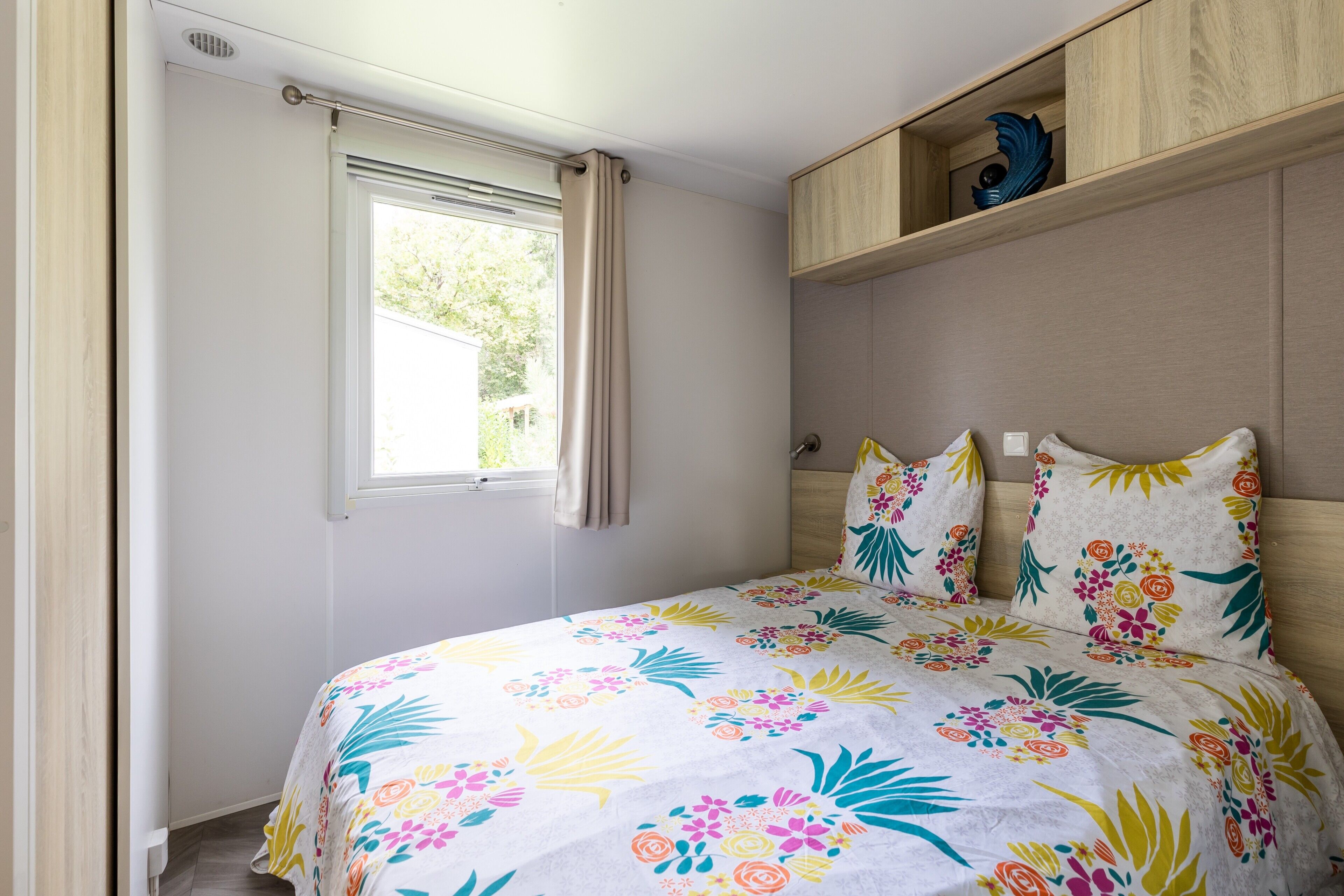 A bedroom with a bed, a window with curtains, a wooden shelf with decorative items, and a headboard.
