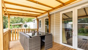 Eine überdachte Terrasse mit einem Tisch aus Glasplatten und Korbstühlen, Holzboden und Blick auf ein Haus und einen Garten.