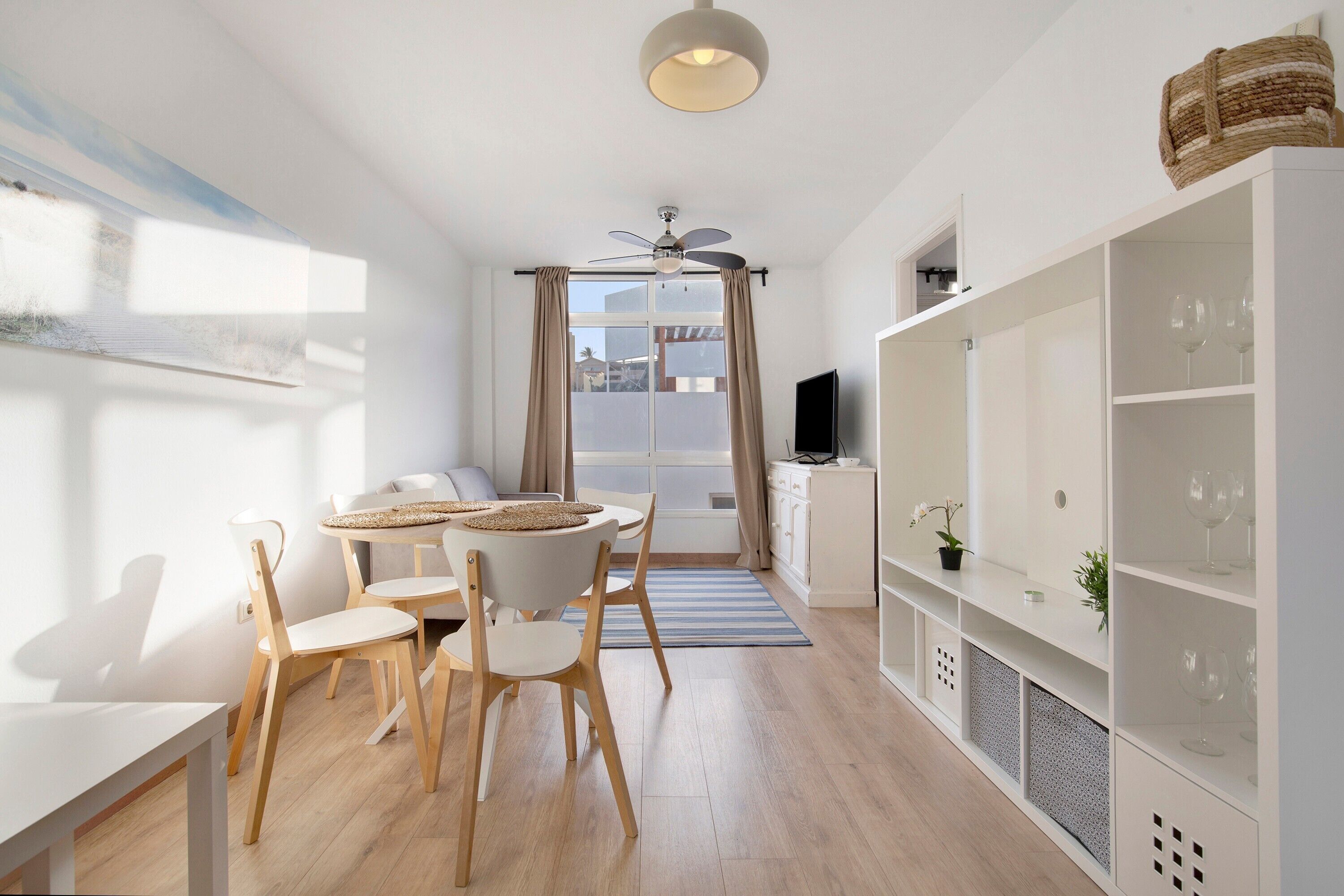 A modern living room with a dining area, a ceiling fan, a TV, and a bookshelf.