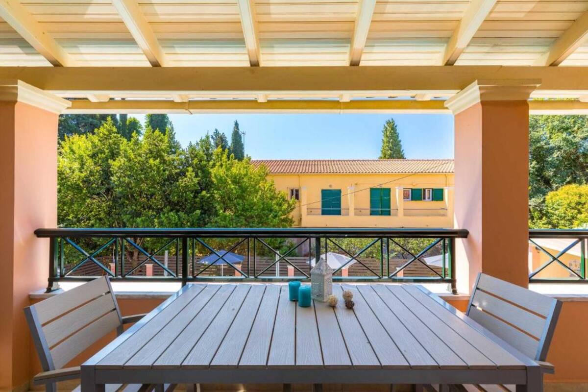 A covered outdoor seating area with a table and chairs, overlooking a building with a tiled roof and green shutters.