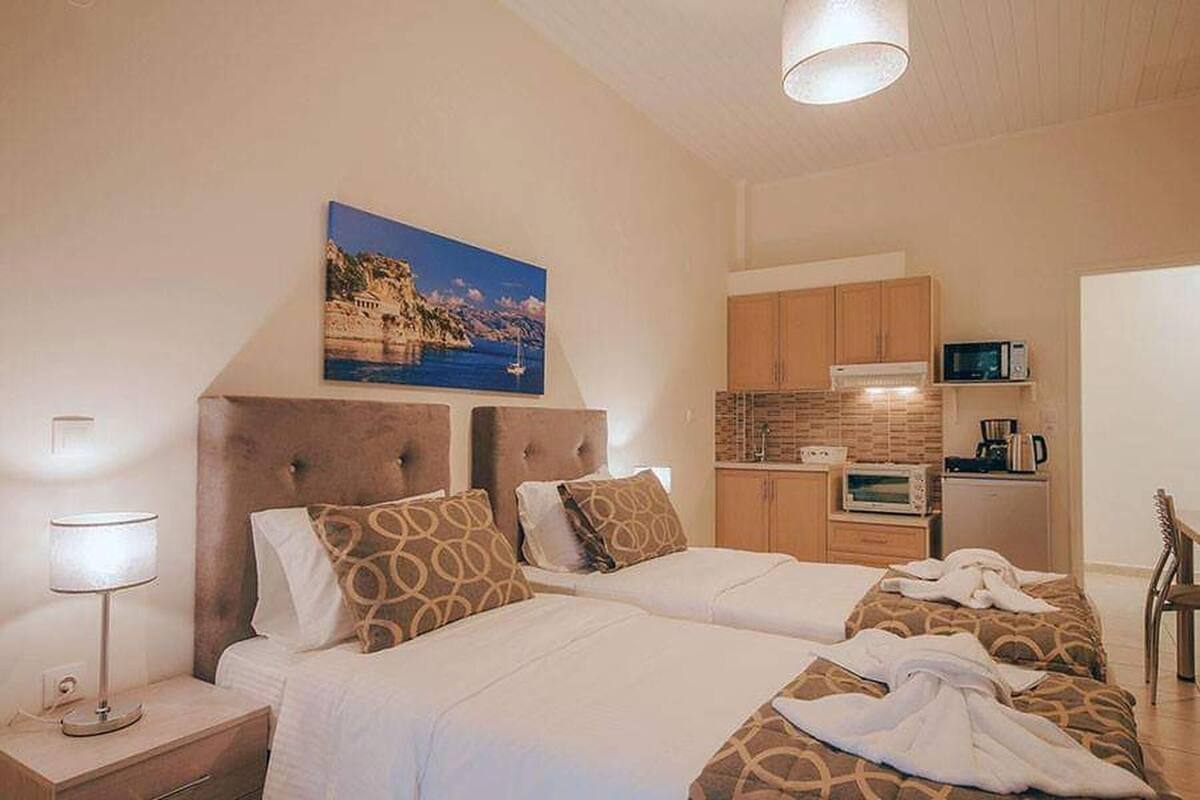 A hotel room with two beds, a kitchenette, a painting of a coastal scene, and a lamp.