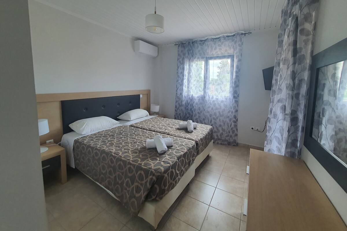 A hotel room with two beds, a wooden headboard, a window with curtains, a bedside table with a lamp, and a flat-screen TV mounted on the wall.