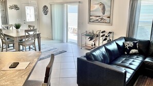 Uma sala de estar com um sofá de couro preto, uma mesa de jantar com cadeiras, um lustre e um quadro de um pássaro.