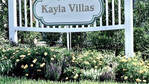 Placa indicando Kayla Villas, com um muro branco e um jardim exuberante ao primeiro plano.