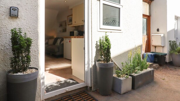 A cozy cottage with a sign reading "Quayside Cottage," a small kitchen, a dining area, and a front door leading to a porch with potted plants.