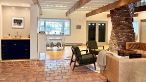A spacious living area with a brick fireplace, a dining area with a large window, and a kitchen with a modern sink and cabinets. - Luxe Cape Cod Farmhouse. Walk to Ellis Landing Beach! (Brewster)