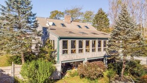 A two-story house with a sloped roof, large windows, and a covered porch. - Luxe Cape Cod Farmhouse. Walk to Ellis Landing Beach! (Brewster)