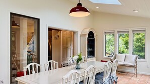 A dining area with a white table and chairs, a red pendant light, and a view of greenery through large windows. - Luxe Cape Cod Farmhouse. Walk to Ellis Landing Beach! (Brewster)