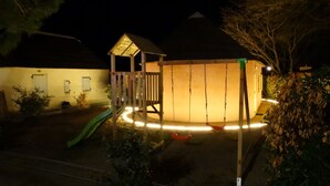 A wooden play structure with a slide and swings, surrounded by a fence and a small house-like building. - Kleines Haus mit Garten, Ideal für Ruhe, Meer, Funboarding und Windsurfing (Le Barcarès)
