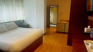 A hotel room with a large bed, a bedside table with a phone, a wooden floor, and a small table with a tissue box and a glass of water. - Hotel Aruni Ancol (Jakarta)