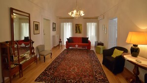 Una sala de estar con un sofá rojo, un sillón negro, una mesa de centro de madera, una mesa de centro de vidrio, un tapete estampado y una araña.