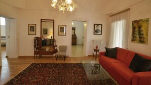 Un salón con un sofá rojo, una mesa de centro de vidrio, un tapete estampado, una araña y cuadros enmarcados en las paredes.