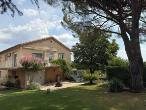 A two-story house with a balcony, surrounded by a well-maintained garden and a large tree.