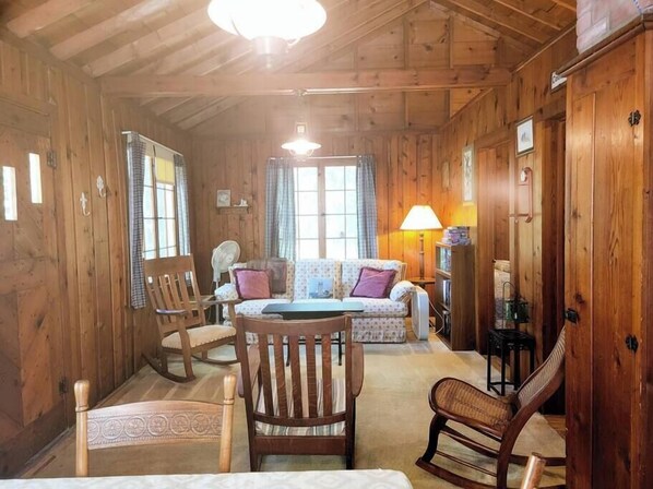 A cozy cabin interior with wooden walls, a sofa, two rocking chairs, a table, and a lamp.