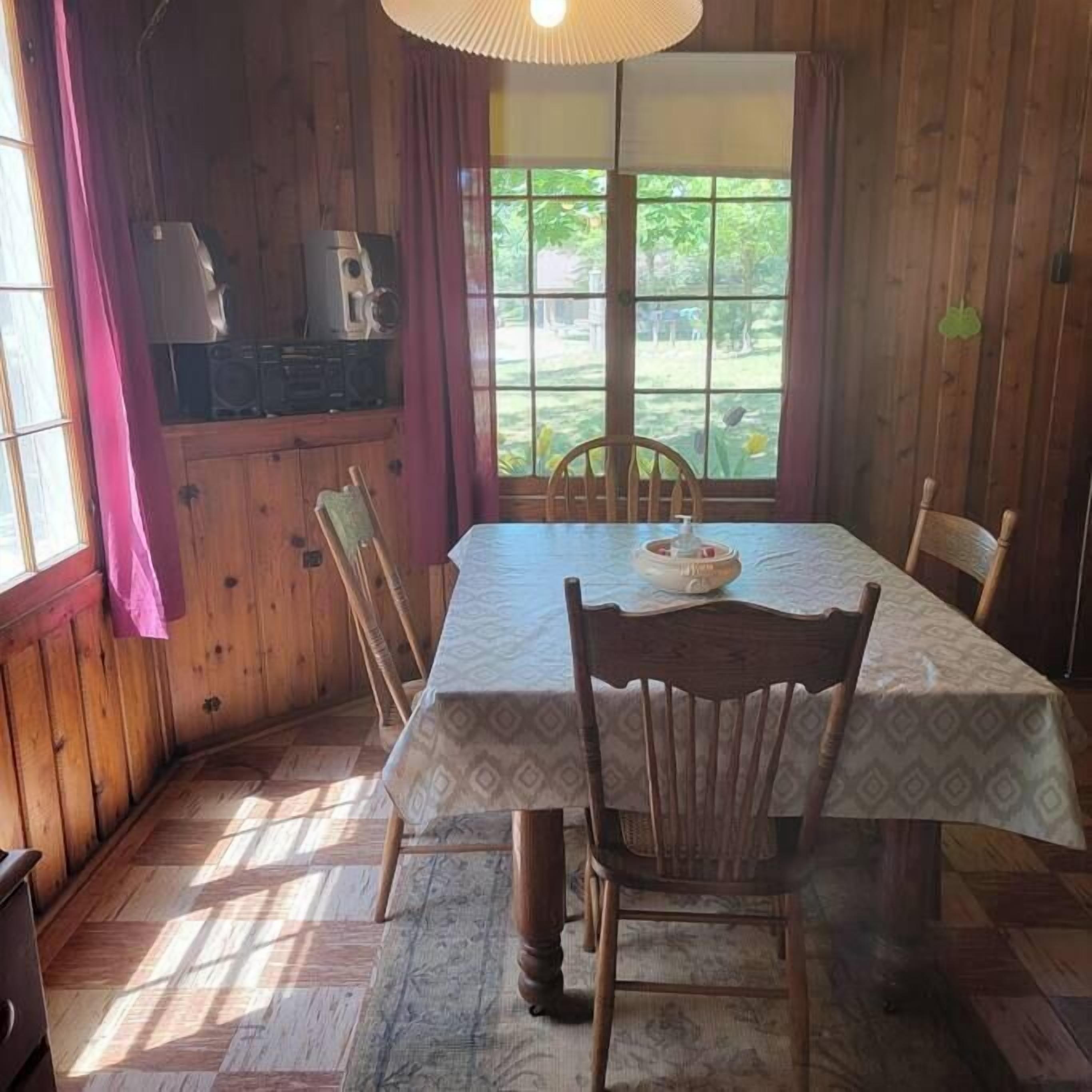 A wooden dining room with a table set for four, a window with curtains, and a view of greenery outside.