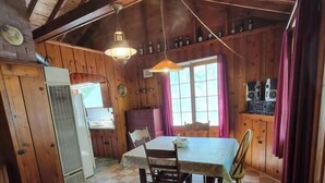 Een knus houten interieur met een eettafel, stoelen, een zwart-wit geruite vloer, een keukenhoek met kastjes en een koelkast, en een raam met gordijnen.