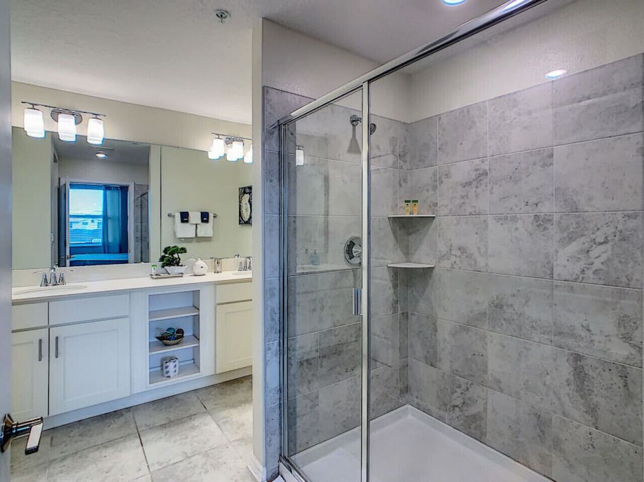 Une salle de bain moderne dotée d’une douche à parois de verre, d’un grand miroir et d’un meuble-lavabo blanc avec un évier et un plan de travail.