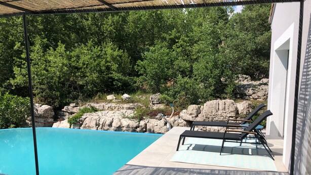 Un espace piscine avec une eau d’un bleu limpide, un patio couvert doté de chaises longues et un paysage rocheux en arrière-plan.