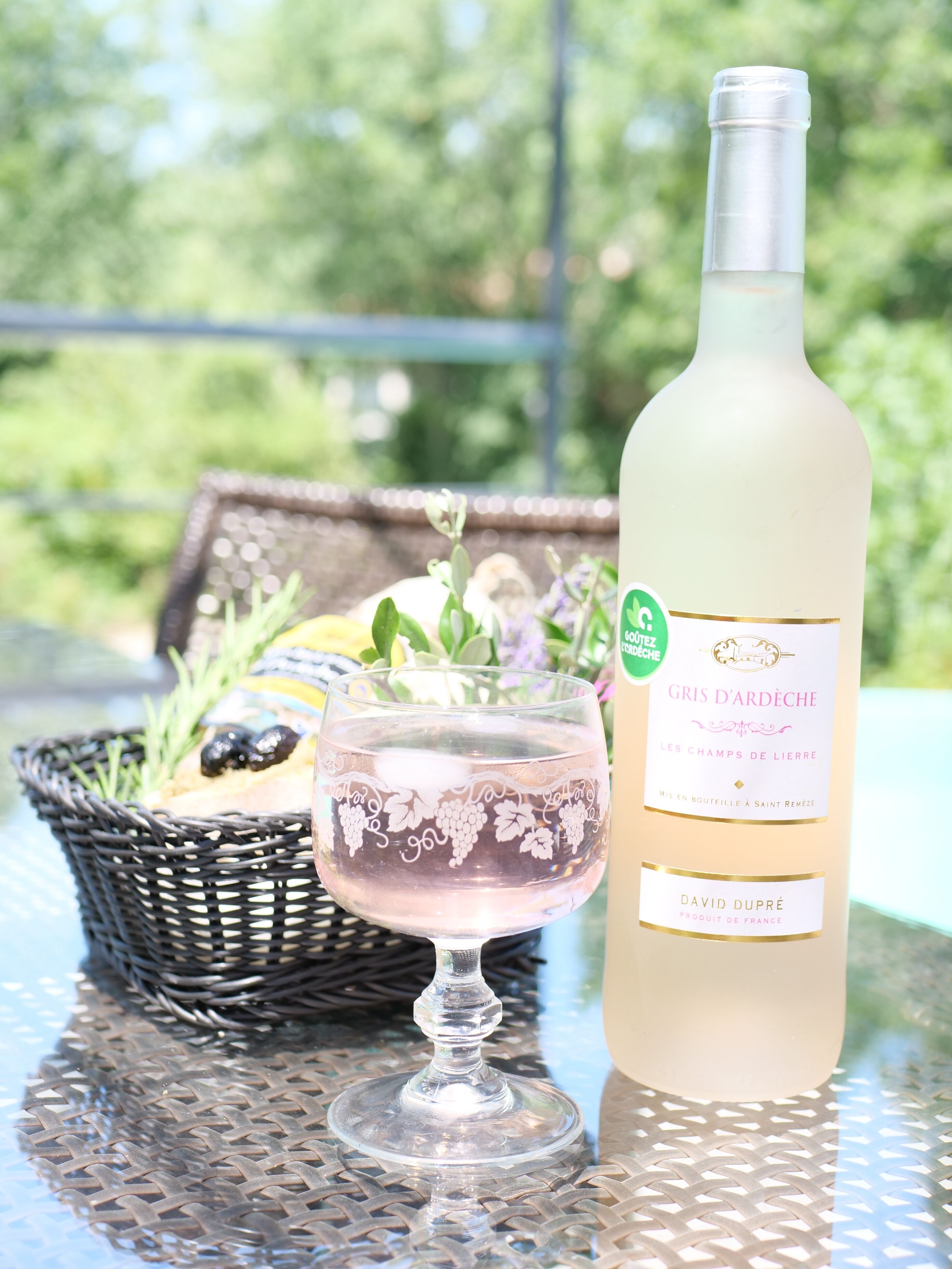 Une bouteille de vin Gris d'Ardèche, un verre de vin rosé et un panier contenant du pain et des herbes aromatiques, le tout posé sur une table en métal à l'extérieur.