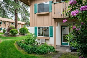 A two-story building with a garden, a balcony with purple flowers, and a window with a view of the garden.