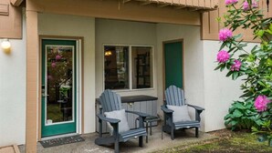 Ein überdachter Außenbereich mit zwei Adirondack-Stühlen, einem Tisch, einer grünen Tür und einem Fenster.