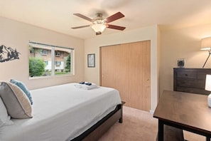 Un dormitorio con cama, ventilador de techo, ventana con vistas, puerta de madera y una mesita.