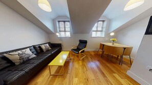 A modern living room with a black leather sofa, a wooden coffee table, a black leather chair, a wooden dining table with chairs, and two windows with blinds.