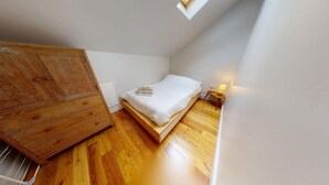 Ein kleiner, gut beleuchteter Raum mit Holzboden, einem Bett mit weißer Bettwäsche, einem Holzschrank und einem kleinen Nachttisch mit Lampe.