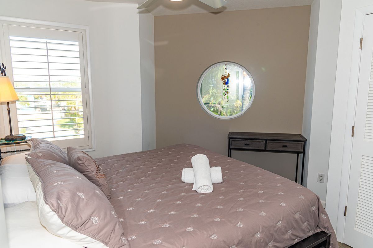 Un lit bien fait, avec une couvre-lit rose et des oreillers blancs, un miroir rond accroché au mur et une fenêtre avec des stores.