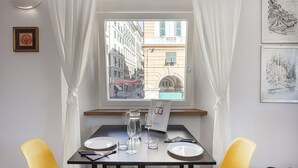 Uma área de refeições com uma mesa posta para dois, uma janela com vista para a rua e um quadro na parede.