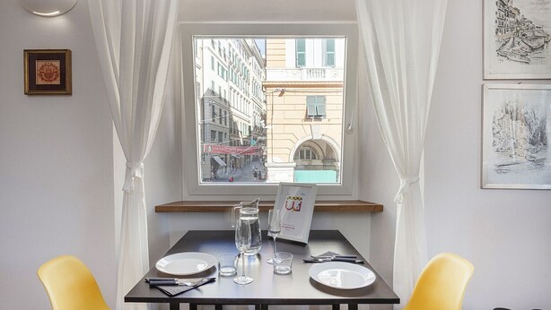 A dining area with a table set for two, a window with a view of a city street, and a framed picture on the wall. - San Lorenzo View Apartment 4 by Wonderful Italy (Genova)