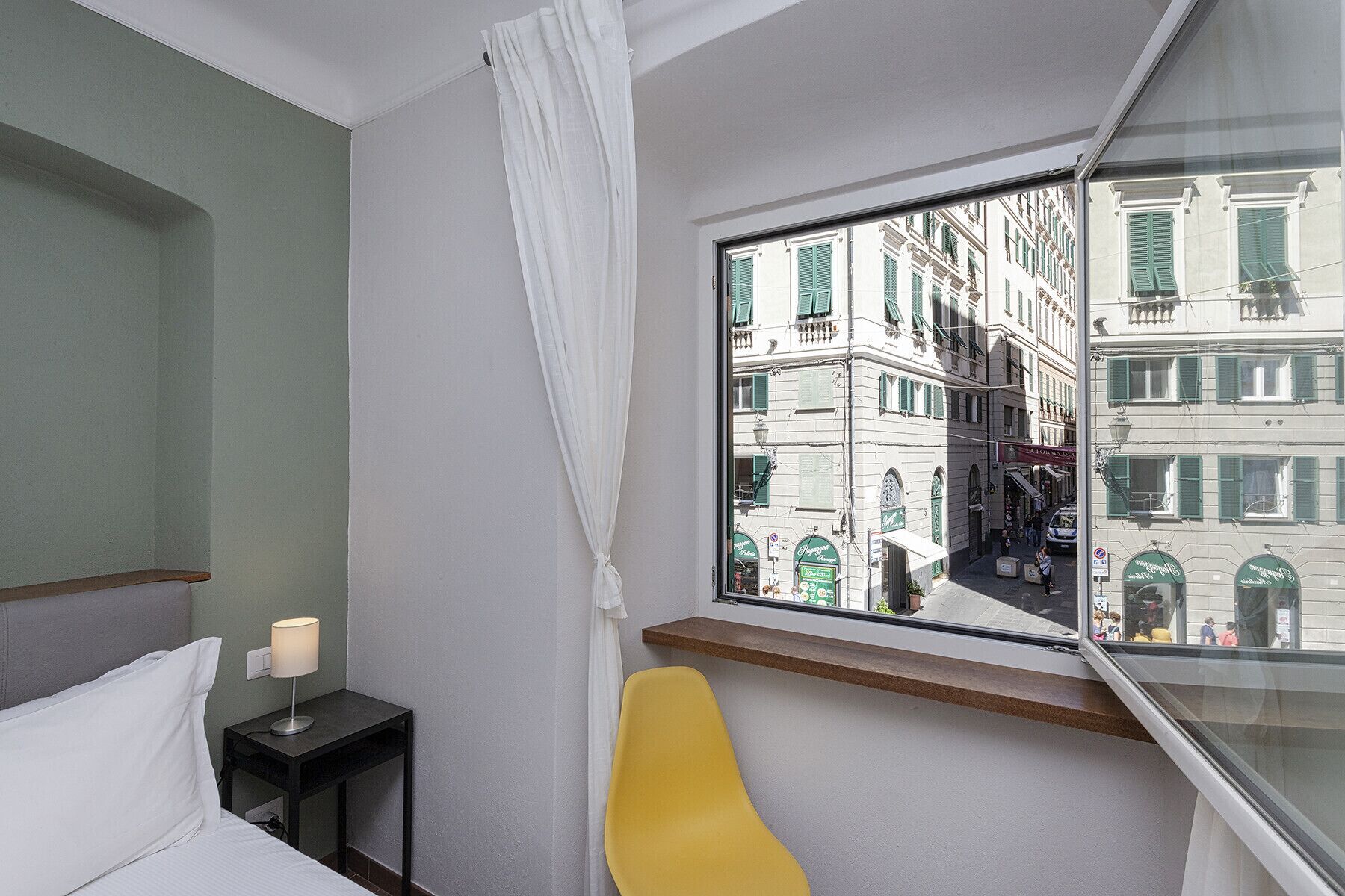 A hotel room with a bed, a nightstand with a lamp, a yellow chair, and a window with a view of buildings.