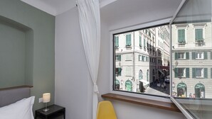 Quarto de hotel com cama, mesa de cabeceira com abajur, cadeira amarela e janela com vista para edifícios.