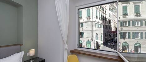 A hotel room with a bed, a nightstand with a lamp, a yellow chair, and a window with a view of buildings.