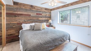 A bedroom with a wooden wall, a bed with a gray quilt, a ceiling fan, and a window with a view of trees. - Riverwalk Retreat | Private Deck | Walk to Main St (Ouray)