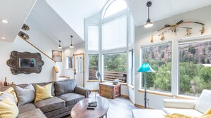A spacious living room with a high ceiling, large windows offering a view of a forested area, a cozy sofa set, and a small wooden table. - Riverwalk Retreat | Private Deck | Walk to Main St (Ouray)