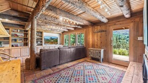 A rustic cabin interior with exposed wooden beams, a leather sofa, a large window with a scenic view, and a bookshelf. - Solitude in Sundance - Hot Tub, 4 Bed 4 Bath, Secluded, Views, Quiet, Forested (Sundance)