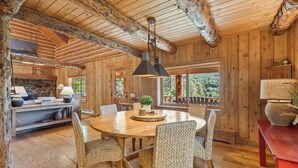 A rustic wooden interior with a round dining table surrounded by woven chairs, a stone fireplace, and a large window offering a view of the outdoors. - Solitude in Sundance - Hot Tub, 4 Bed 4 Bath, Secluded, Views, Quiet, Forested (Sundance)