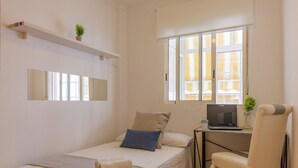 Une petite chambre à coucher, bien rangée, avec un lit, un bureau sur lequel se trouve un ordinateur portable, une chaise, une fenêtre avec des rideaux et une étagère avec une plante.