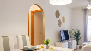 Un coin repas moderne avec une table blanche dressée pour deux personnes, un téléviseur posé sur un meuble et une plante en pot.
